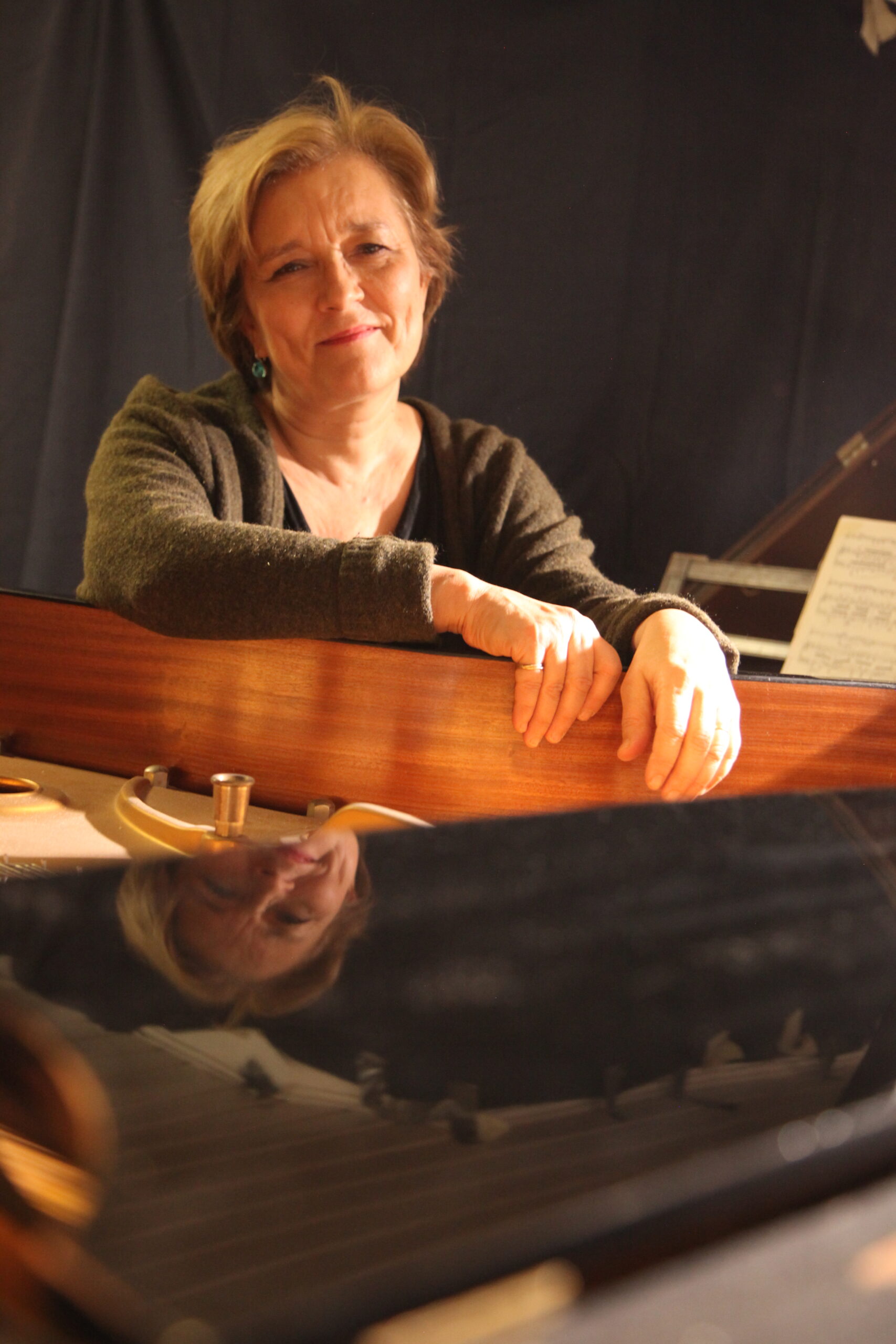 Woman with short red hair in a brown sweater sitting with hands over the edge of a grand piano with a harpsichord in the background.